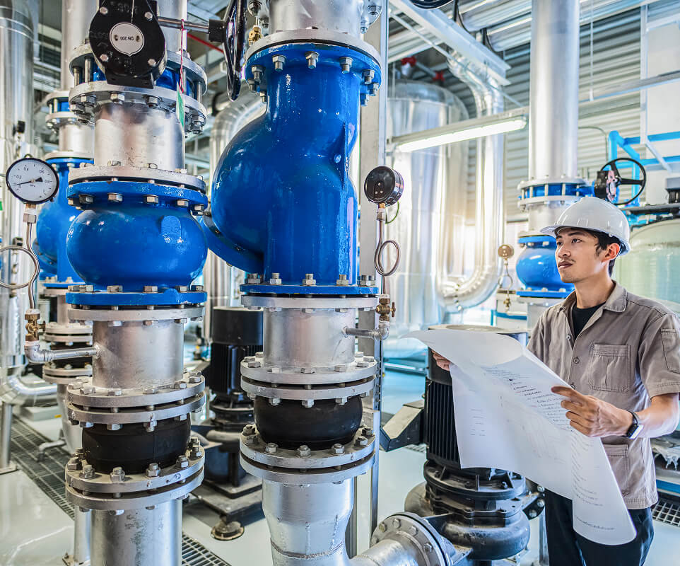 A male engineer wearing a hard hat and holding a blueprint, stands in an industrial facility with large pipes and machinery.